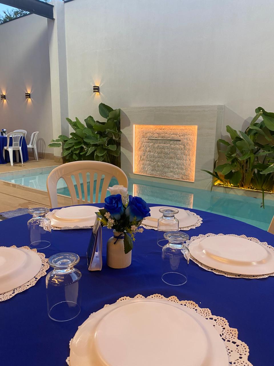 Mesa decorada em azul ao lado da piscina com cascata iluminada e plantas